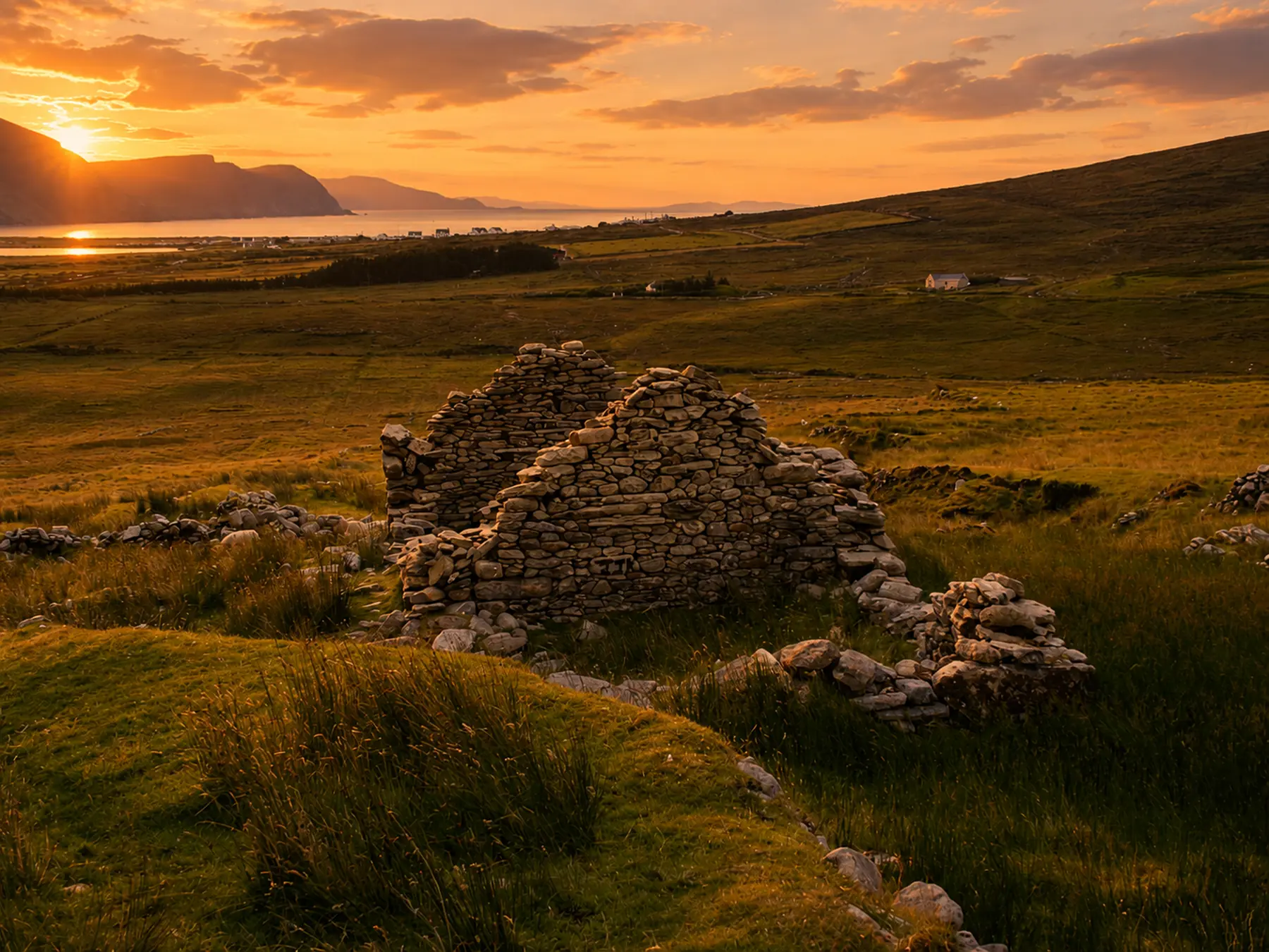 The stone ruins of an old cottage sit in a vast, grassy green valley in Ireland during a vibrant golden sunset, with mountains and the ocean visible in the background.